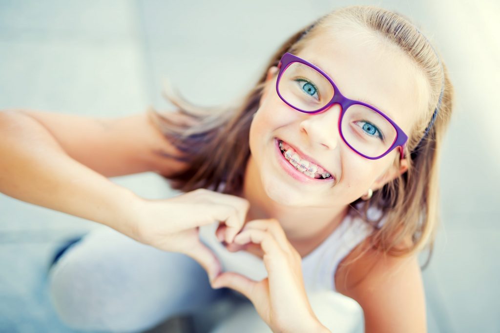 Smiling pre-teen with braces wearing purple glasses with her hands in the shape of a heart.